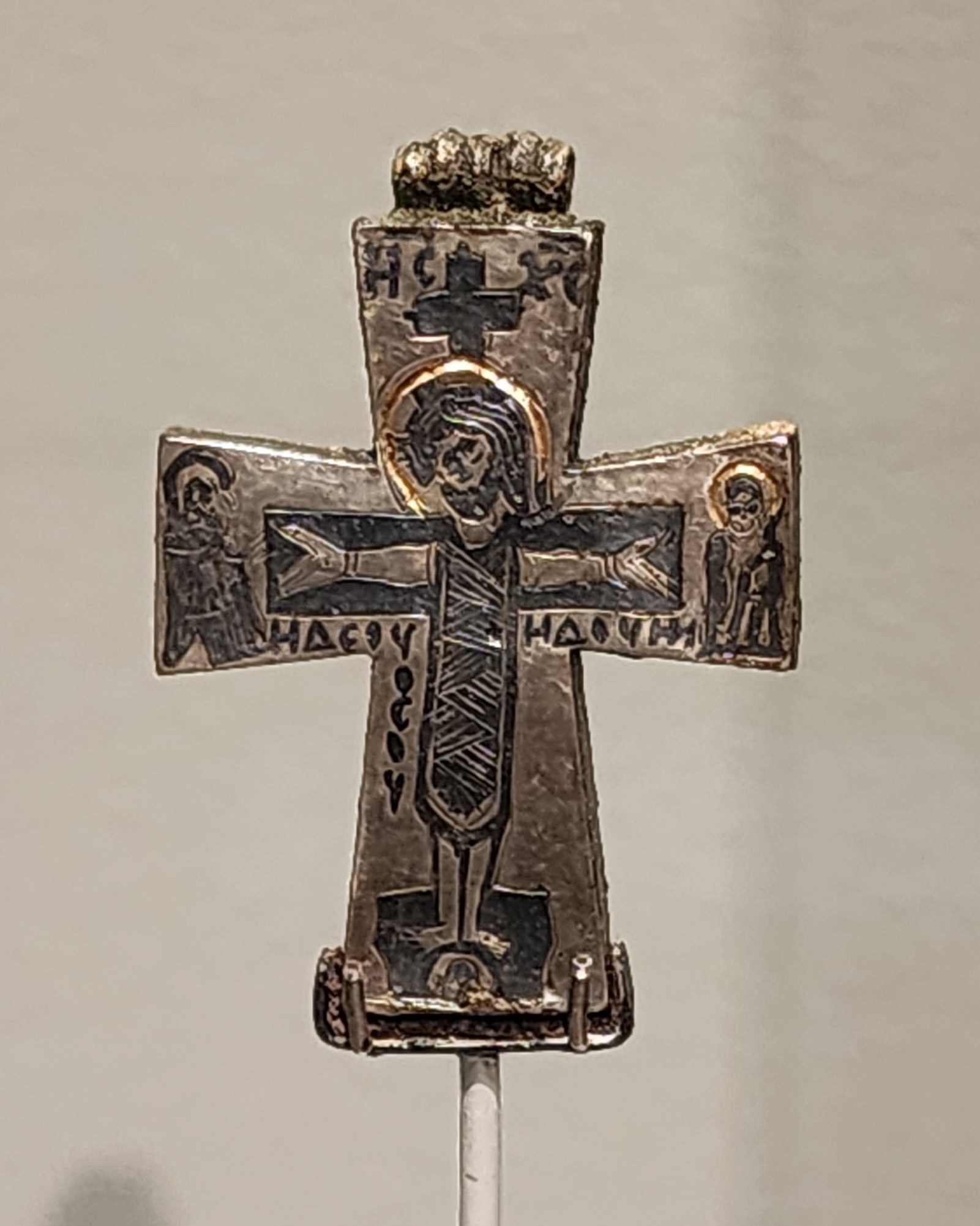 small silver reliquary in the shape of a cross with a crucified Jesus and Greek script written in black
